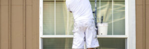 Painter man in white clothes with a protective scarf on his head paints a wooden window trim with a brush while standing on a high ladder leaning against the facade of a residential building